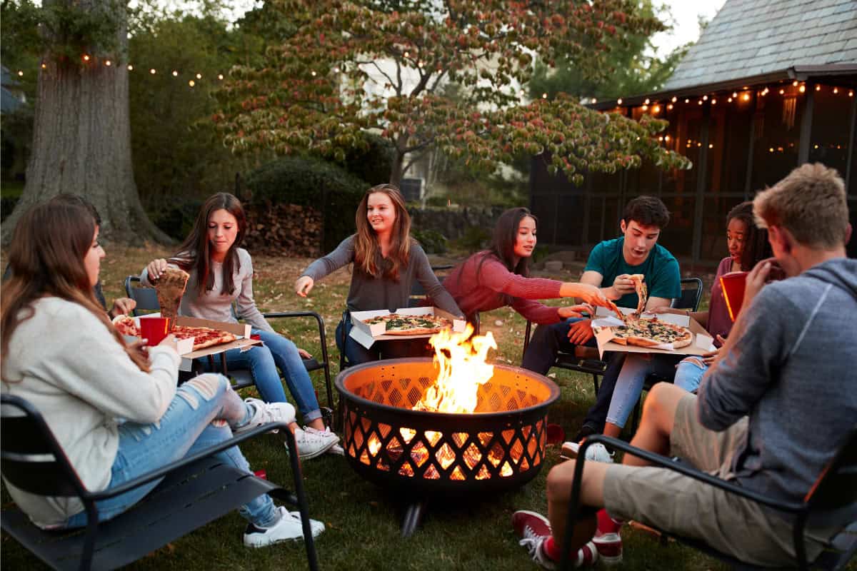 Image of a group of teenagers eating pizza around a burning fire pit.