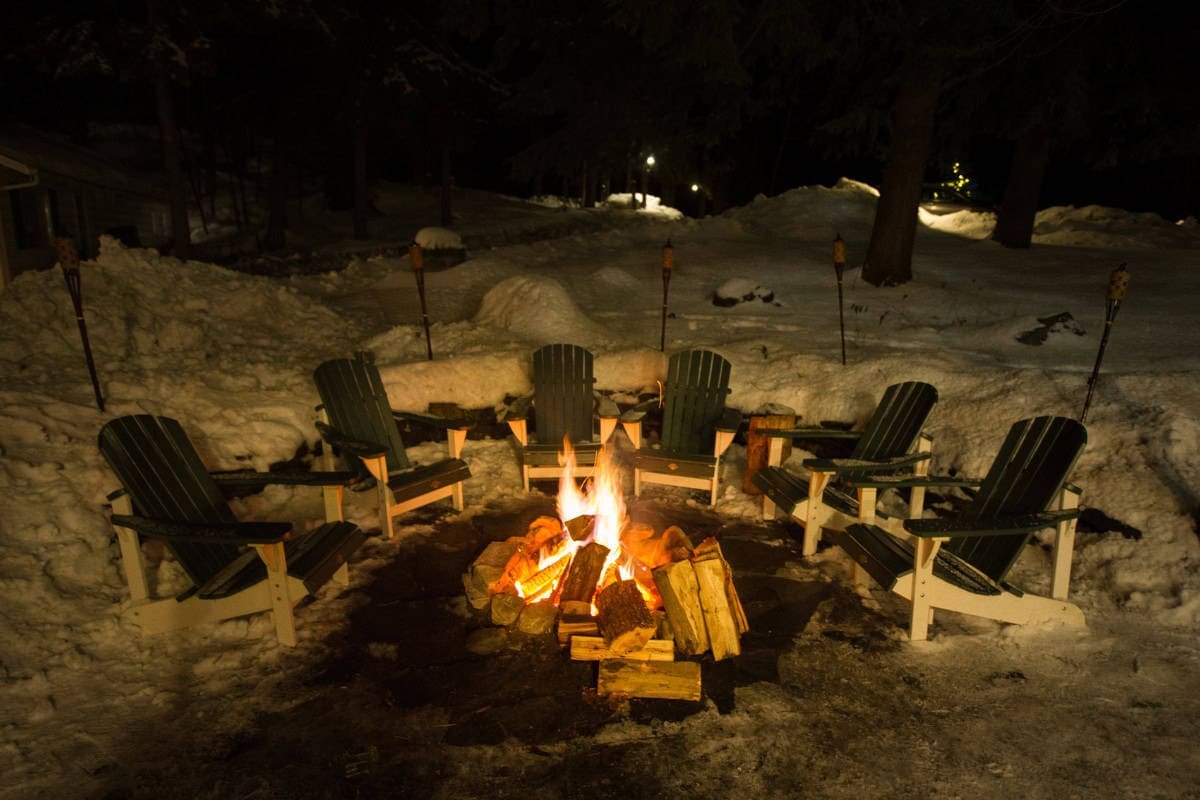 Image of a burning fire pit surrounded by Adirondack chairs after a heavy snowfall.