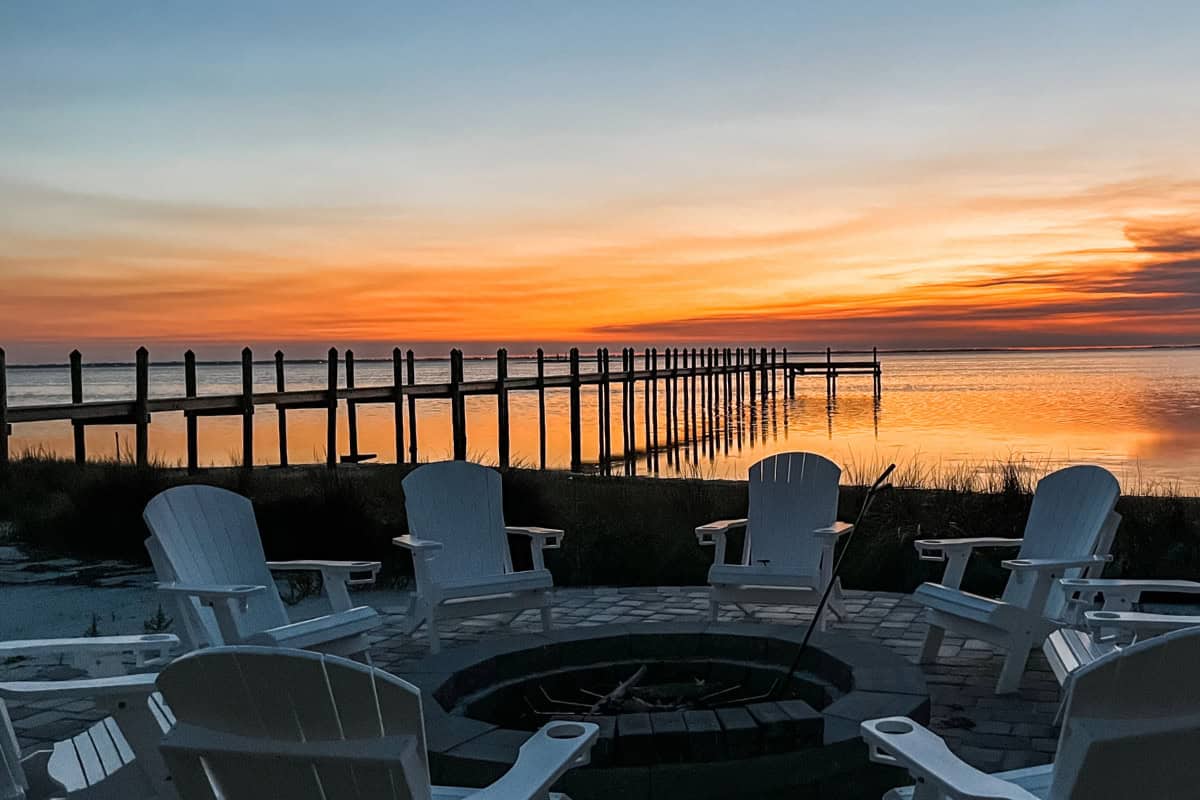 Image of a fire pit and chairs by a river at sunset.