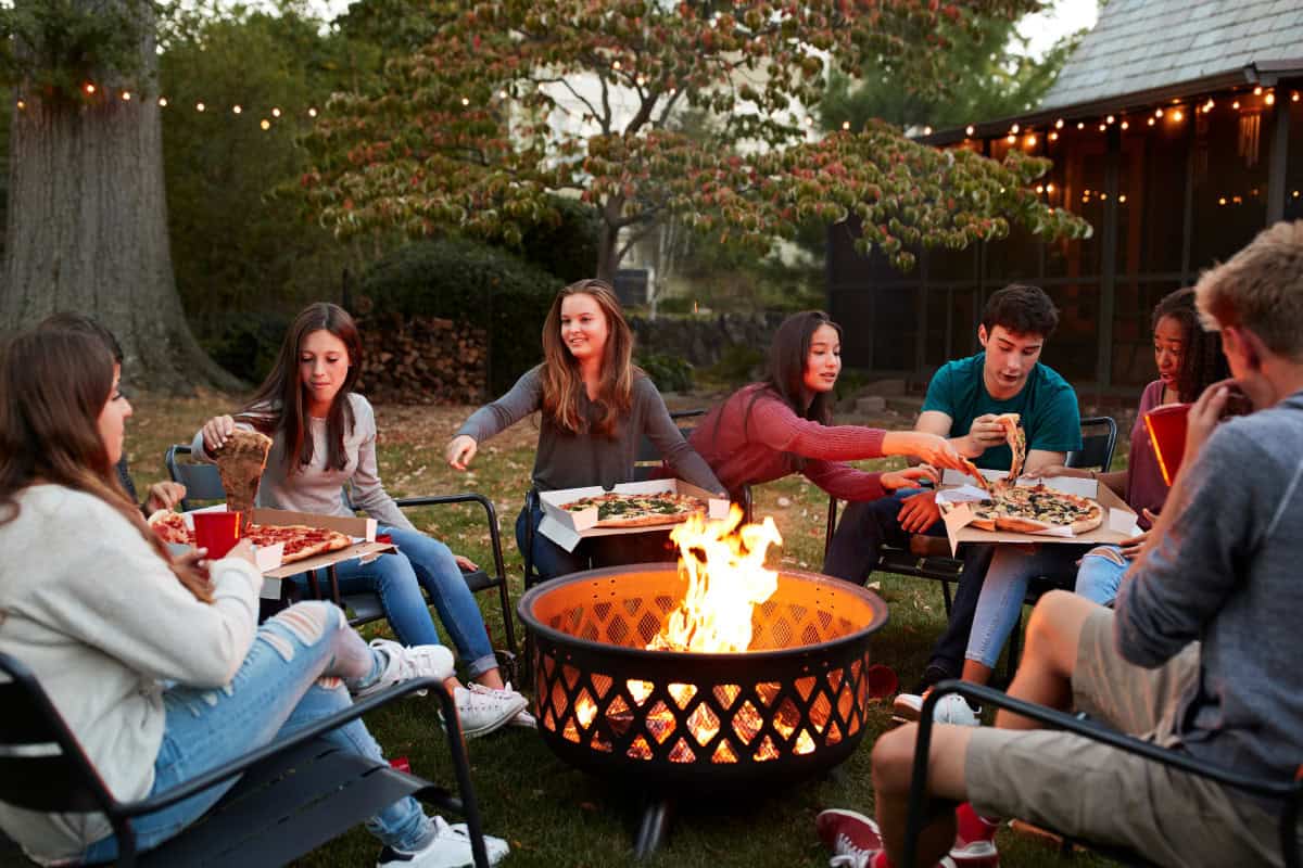 Wood-burning fire pit at the center of a pizza party Image of a group of teenagers eating pizza around a wood-burning fire pit.