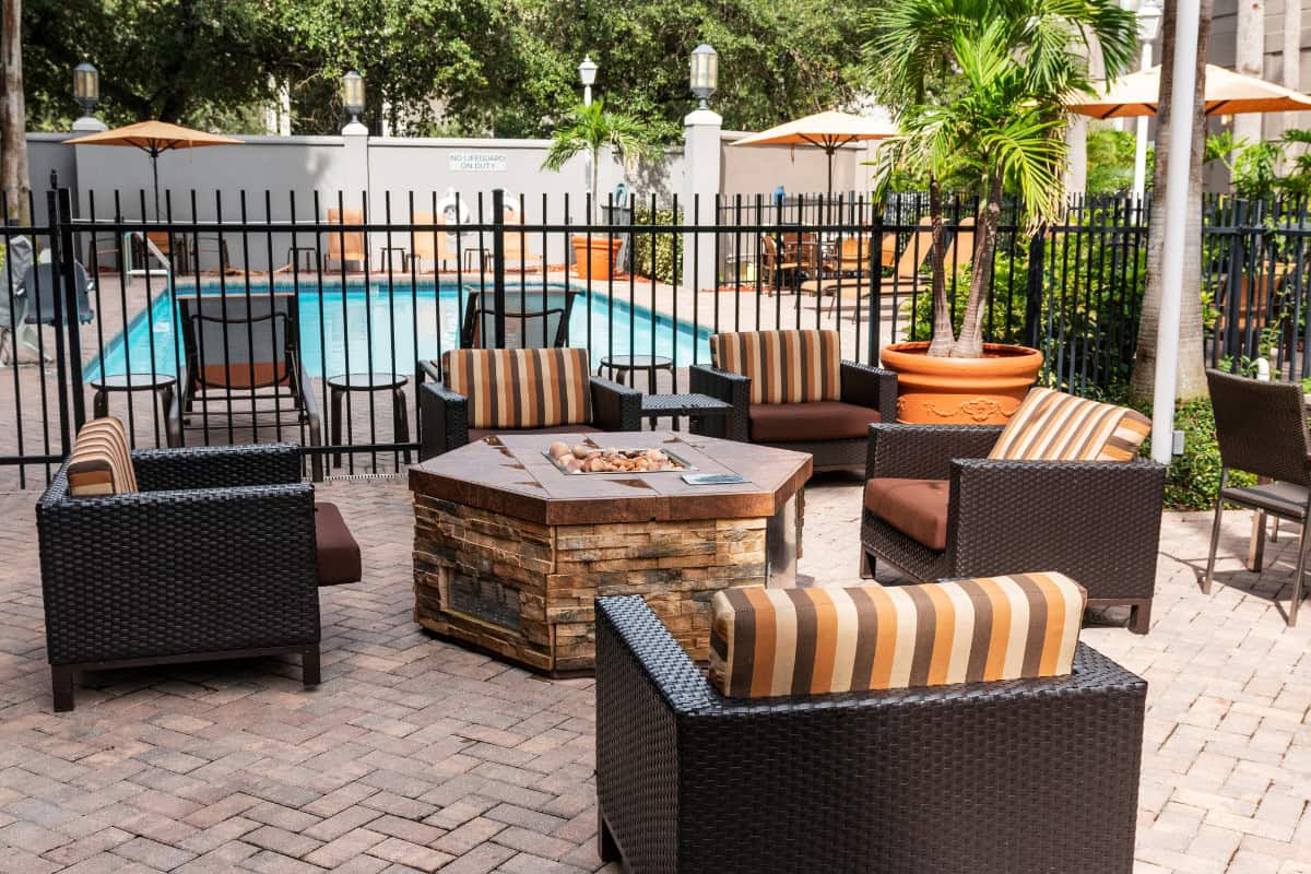 Cozy outdoor seating area with wicker chairs, striped cushions, and a fire pit near a swimming pool, surrounded by lush tropical plants and umbrellas at Backyard Toasty.