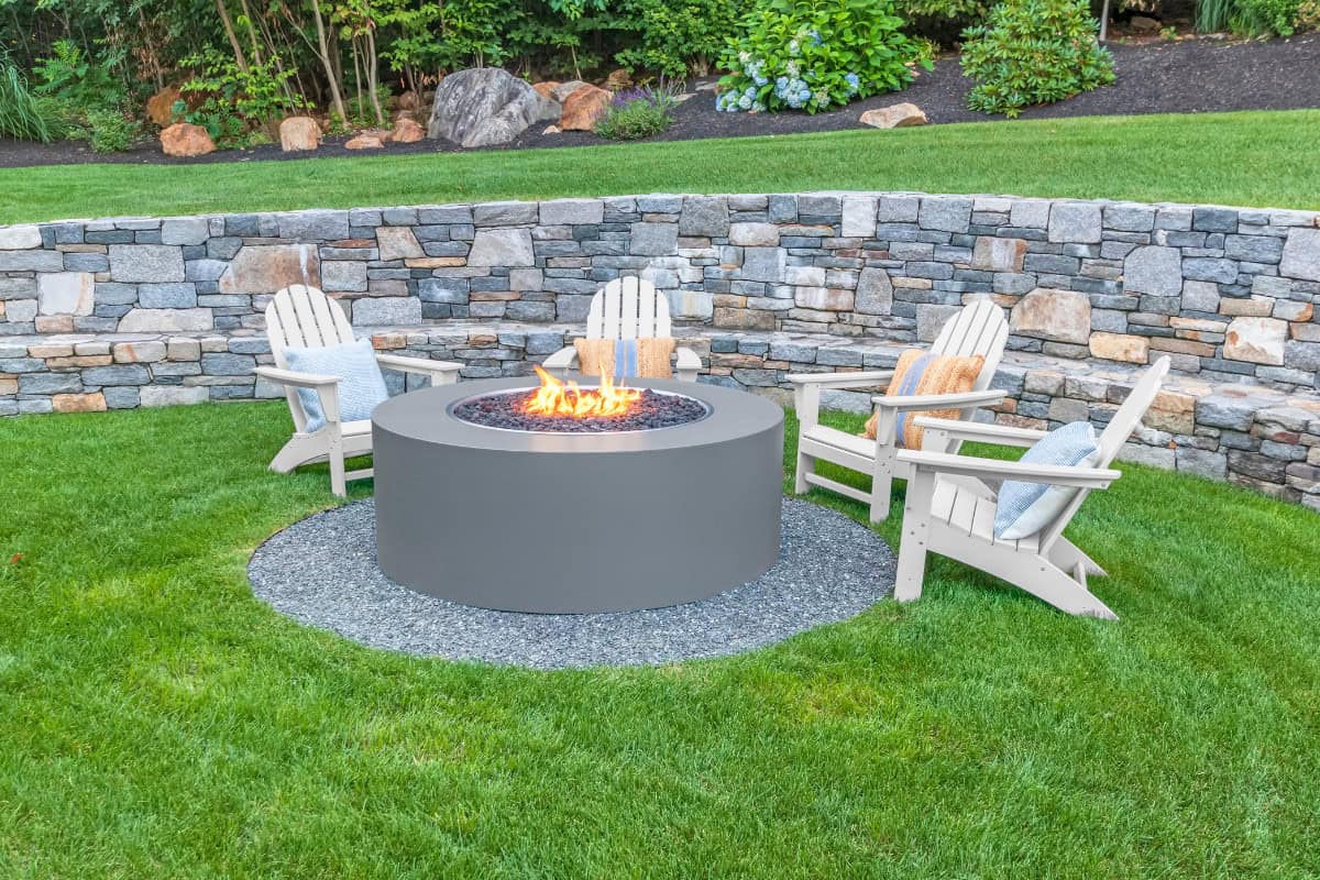 Fire pit outdoor backyard with seating area - Backyard Toasty fire pit setup featuring Adirondack chairs in a landscaped garden with a stone retaining wall and lush green grass.