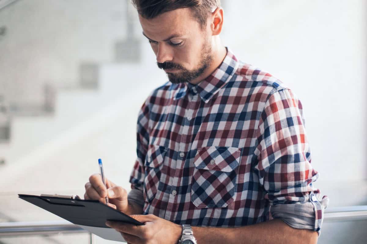 Man writing on clipboard with a pen indoors, casual checklists or planning for backyard design and outdoor projects.